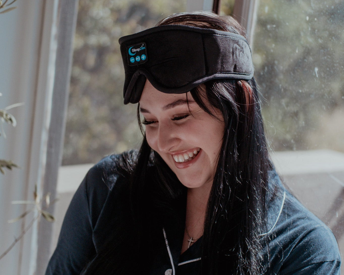 Woman wearing a sleep mask and holding a mug, sitting by a window with a blurred outdoor background.